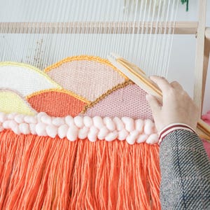 May include: A wooden weaving loom holding a partially completed tapestry with a landscape design. The textile features curved sections in yellow, peach, and orange yarn, topped with a row of thick, pale pink braided wool and long orange fringe.