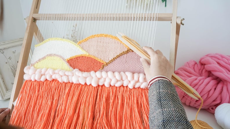 May include: A wooden weaving loom holding a partially completed tapestry with a landscape design. The textile features curved sections in yellow, peach, and orange yarn, topped with a row of thick, pale pink braided wool and long orange fringe.