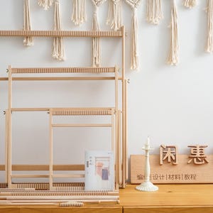 May include: Three light-colored wooden weaving looms of varying sizes arranged in a nested formation. The set includes several wooden rods and a small comb tool. A wooden sign with the text 闲惠居家 and sheweave is placed to the right.