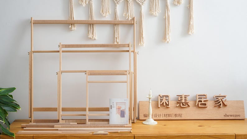 May include: Three light-colored wooden weaving looms of varying sizes arranged in a nested formation. The set includes several wooden rods and a small comb tool. A wooden sign with the text 闲惠居家 and sheweave is placed to the right.