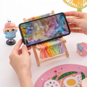 May include: A wooden phone stand with a rainbow macrame design. The phone displays a colorful floral image. The stand has a small, decorative fringe and is held by hands. A small figurine is on the left.