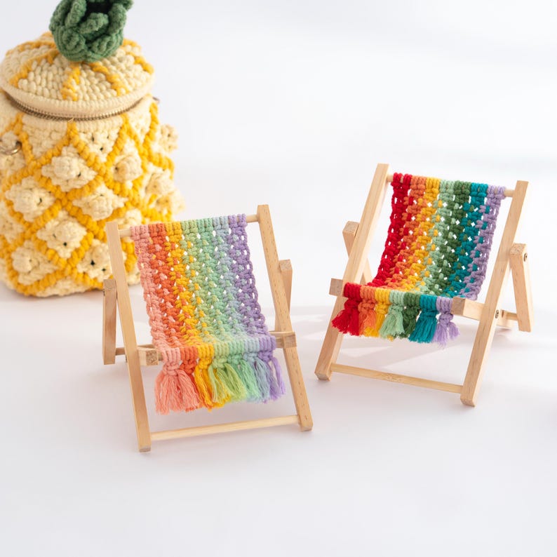 May include: Two miniature wooden beach chairs with rainbow-colored macrame seats and tassels. A yellow pineapple-shaped crochet bag with a green top is in the background. The chairs are set against a white background.