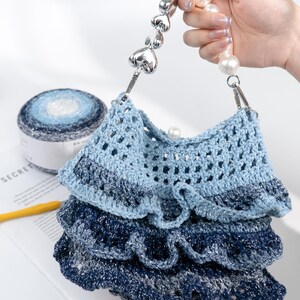 May include: A handmade blue crochet bag with a layered design. The bag features a silver heart-shaped handle and pearl accents. A ball of blue and white yarn is visible in the background, suggesting a craft theme.