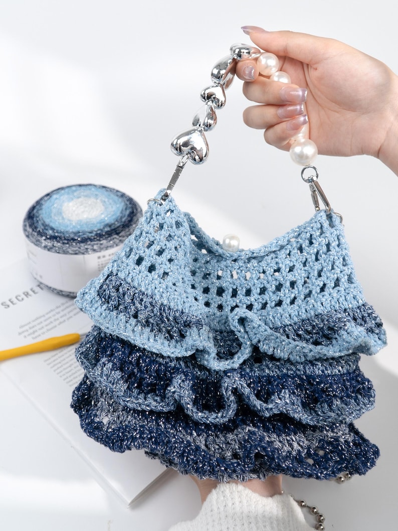 May include: A handmade blue crochet bag with a layered design. The bag features a silver heart-shaped handle and pearl accents. A ball of blue and white yarn is visible in the background, suggesting a craft theme.