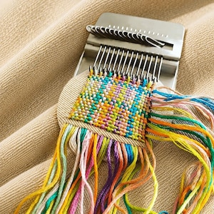 May include: A metal darning loom tool with a series of vertical hooks, currently holding a square patch of woven fabric. The weave features a multicolored pattern of blue, yellow, orange, pink, and green threads, with loose yarn ends extending outward.