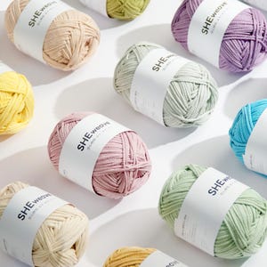 Puede incluir: Una variedad de ovillos de lana coloridos, incluyendo tonos de rosa, verde, amarillo y azul, están dispuestos sobre una superficie blanca. Cada ovillo tiene una etiqueta blanca con el texto "SHE weave". La lana parece suave y bien enrollada, ideal para tejer o hacer ganchillo.