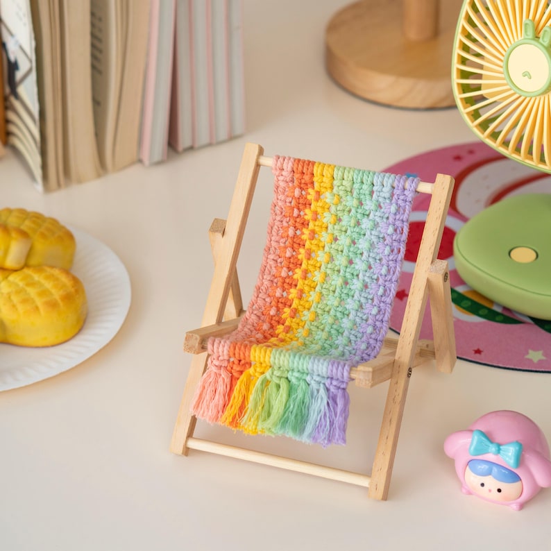 May include: A miniature wooden beach chair with a rainbow-colored crocheted seat and fringe. The chair is made of light-colored wood and has a colorful, textured seat. A small pink figurine is in the foreground.