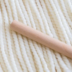 May include: A smooth, cylindrical light-colored wooden dowel rests diagonally across a series of parallel, thick, white twisted yarn strands. The yarn features a consistent, rope-like texture, while the wooden rod has a matte finish.