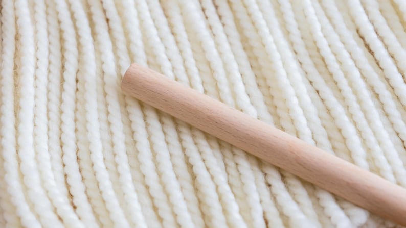 May include: A smooth, cylindrical light-colored wooden dowel rests diagonally across a series of parallel, thick, white twisted yarn strands. The yarn features a consistent, rope-like texture, while the wooden rod has a matte finish.