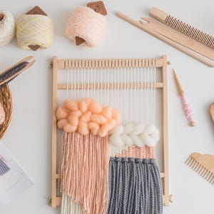 May include: A collection of weaving supplies, including a wooden loom with a partially woven piece in peach, white, and gray.  Also shown are yarn balls, a basket of yarn, weaving tools, and a small wooden tray with scissors and thread.