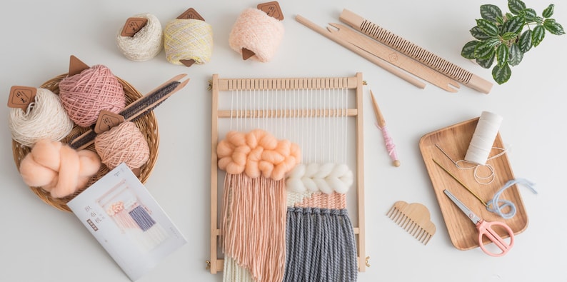 May include: A collection of weaving supplies, including a wooden loom with a partially woven piece in peach, white, and gray.  Also shown are yarn balls, a basket of yarn, weaving tools, and a small wooden tray with scissors and thread.