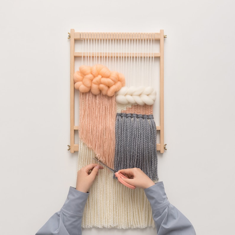 May include: A wooden weaving loom with a partially completed textile art piece. The artwork features peach, cream, and gray yarn in various textures. The hands of a person are working on the weaving with a small tool.