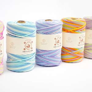 Peut inclure: Cinq bobines de fil multicolores aux tons rose, bleu, violet et orange. Chaque bobine porte une étiquette en papier avec du texte et un logo. Le fil est bien enroulé, montrant les différentes couleurs en rayures.