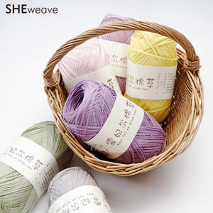 Può includere: Un cestino di vimini pieno di matasse di filato colorato. Il filato è nelle tonalità del lavanda, giallo, rosa chiaro e verde. Ogni matassa ha un'etichetta bianca con testo. Il cestino è realizzato in materiale intrecciato marrone chiaro. Il marchio "SHEweave" è in alto a sinistra.