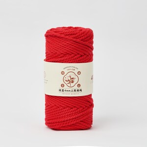 May include: A cylindrical spool of vibrant red cotton cord wrapped in a cream-colored paper band. The band features a central illustration of a cotton plant, the text 100% cotton, and the Chinese characters 闺惠4mm三股棉绳 printed in dark brown.