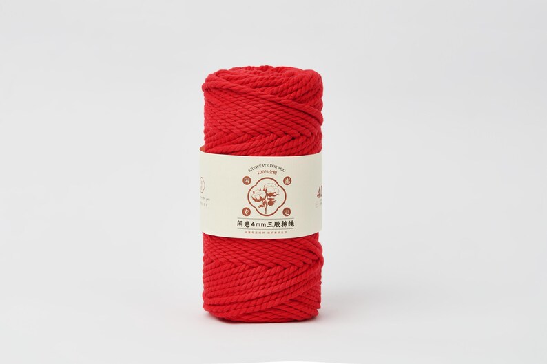 May include: A cylindrical spool of vibrant red cotton cord wrapped in a cream-colored paper band. The band features a central illustration of a cotton plant, the text 100% cotton, and the Chinese characters 闺惠4mm三股棉绳 printed in dark brown.