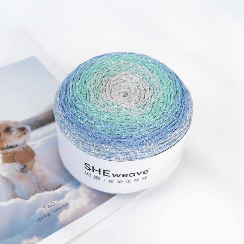 May include: A ball of yarn with a gradient of colors, including blue, green, and silver. The yarn is wrapped around a white label with the text "SHEweave" and Chinese characters. The yarn is sitting on a white surface.