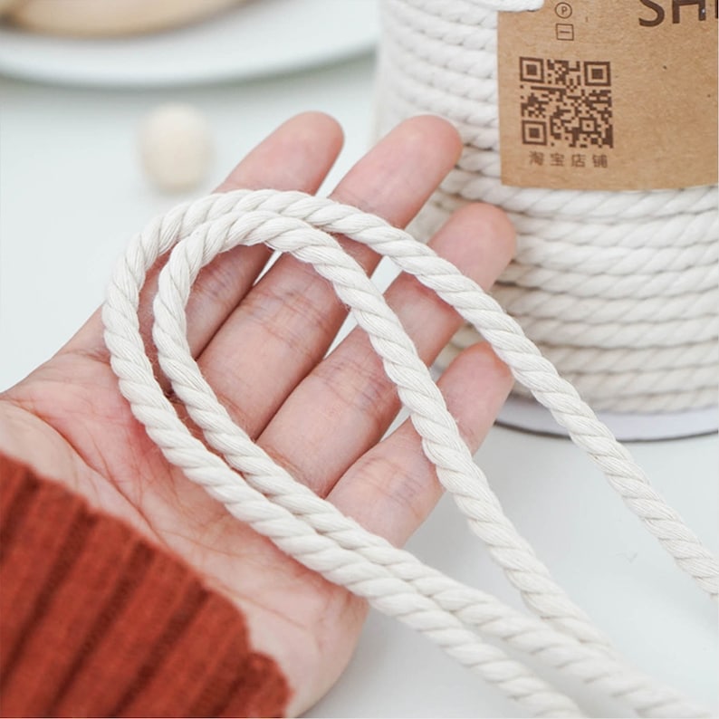 May include: Close-up of off-white cotton rope held in a hand, with a spool of the same rope in the background. The rope is tightly twisted, and the texture is visible. The background is blurred, with a label visible on the spool.