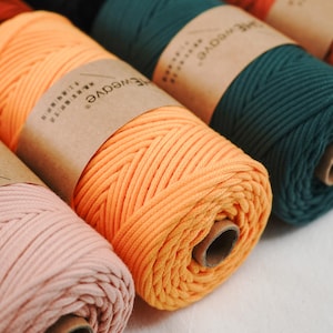 May include: Close-up of several spools of colorful macrame cord. The cord is in shades of orange, green, and pink, with brown paper labels wrapped around each spool. The cord is tightly wound, showing the texture of the material. The image suggests a craft or DIY theme.