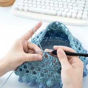 May include: A person crocheting a blue and silver hat. The crochet project is in progress, with the hook and yarn visible. The background includes a white keyboard and a wooden stand.