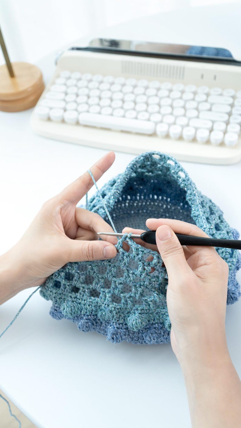 May include: A person crocheting a blue and silver hat. The crochet project is in progress, with the hook and yarn visible. The background includes a white keyboard and a wooden stand.