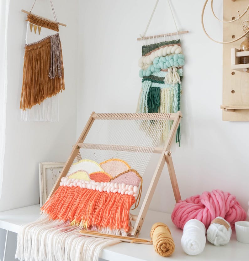 May include: A wooden weaving loom with a colorful woven wall hanging in progress. The artwork features orange, pink, yellow, and white yarn. Two other finished wall hangings are displayed, along with yarn balls and supplies.