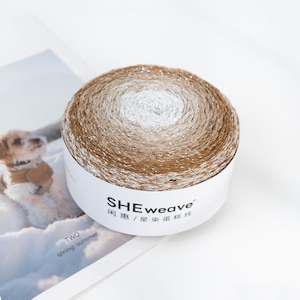 May include: A round skein of yarn with a gradient color scheme, transitioning from white to light brown and dark brown. The yarn is wrapped around a white cardboard tube with the brand name "SHEweave" printed on it. The image is on a white background.