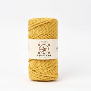 May include: A cylindrical spool of mustard yellow cotton cord wrapped in a paper band. The band features a central graphic of a cotton boll, the text 4mm, and Chinese characters. The cord has a twisted, three-strand construction throughout the spool.