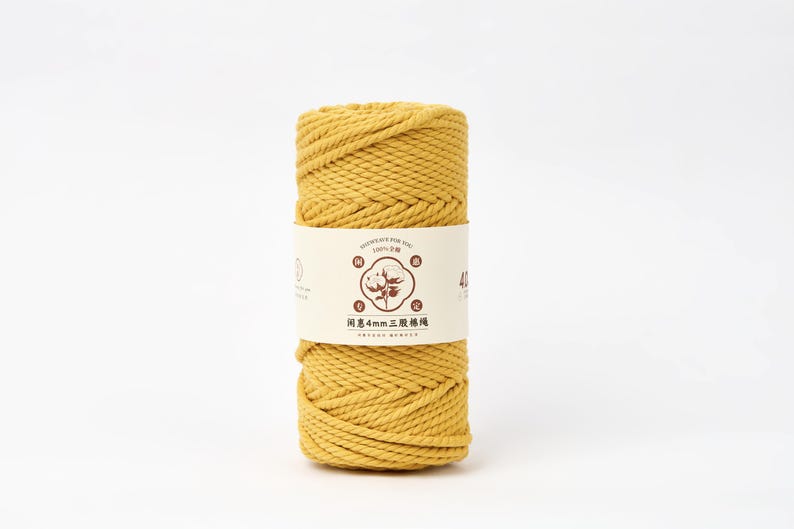 May include: A cylindrical spool of mustard yellow cotton cord wrapped in a paper band. The band features a central graphic of a cotton boll, the text 4mm, and Chinese characters. The cord has a twisted, three-strand construction throughout the spool.