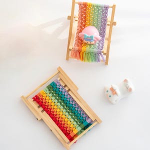 May include: Two miniature wooden beach chairs with rainbow-colored woven seats. One chair has a pink figurine with a blue bow. A white ceramic figurine with a pink bow is also present.