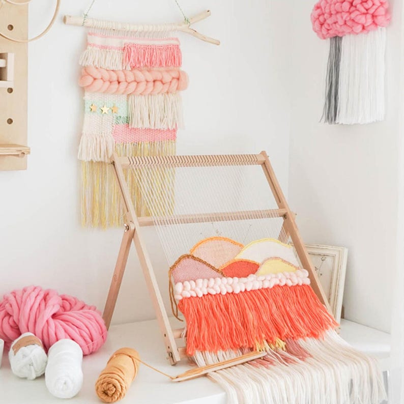 May include: A wooden weaving loom with a colorful woven landscape design. The artwork features orange, yellow, and pink hues. Additional woven wall hangings in pink and white are displayed, along with yarn in various colors.