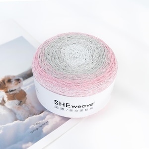 May include: A ball of yarn with a gradient of pink, silver, and gray. The yarn is wrapped around a white label with the text "SHEweave" and Chinese characters. The yarn is on a white surface.