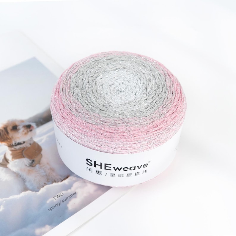 May include: A ball of yarn with a gradient of pink, silver, and gray. The yarn is wrapped around a white label with the text "SHEweave" and Chinese characters. The yarn is on a white surface.