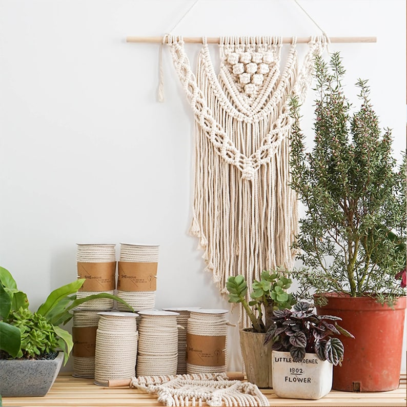 Pu&ograve; includere: Un arazzo macram&egrave; color crema con un design geometrico &egrave; appeso sopra diversi rocchetti di corda color naturale. I rocchetti di corda sono impilati su una superficie di legno, accanto a piante in vaso.