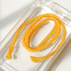 May include: A coiled length of bright yellow twisted rope with a textured surface, resting inside a clear, rectangular plastic container. The rope features a distinct braided construction with visible fibers at the frayed ends of the cord.