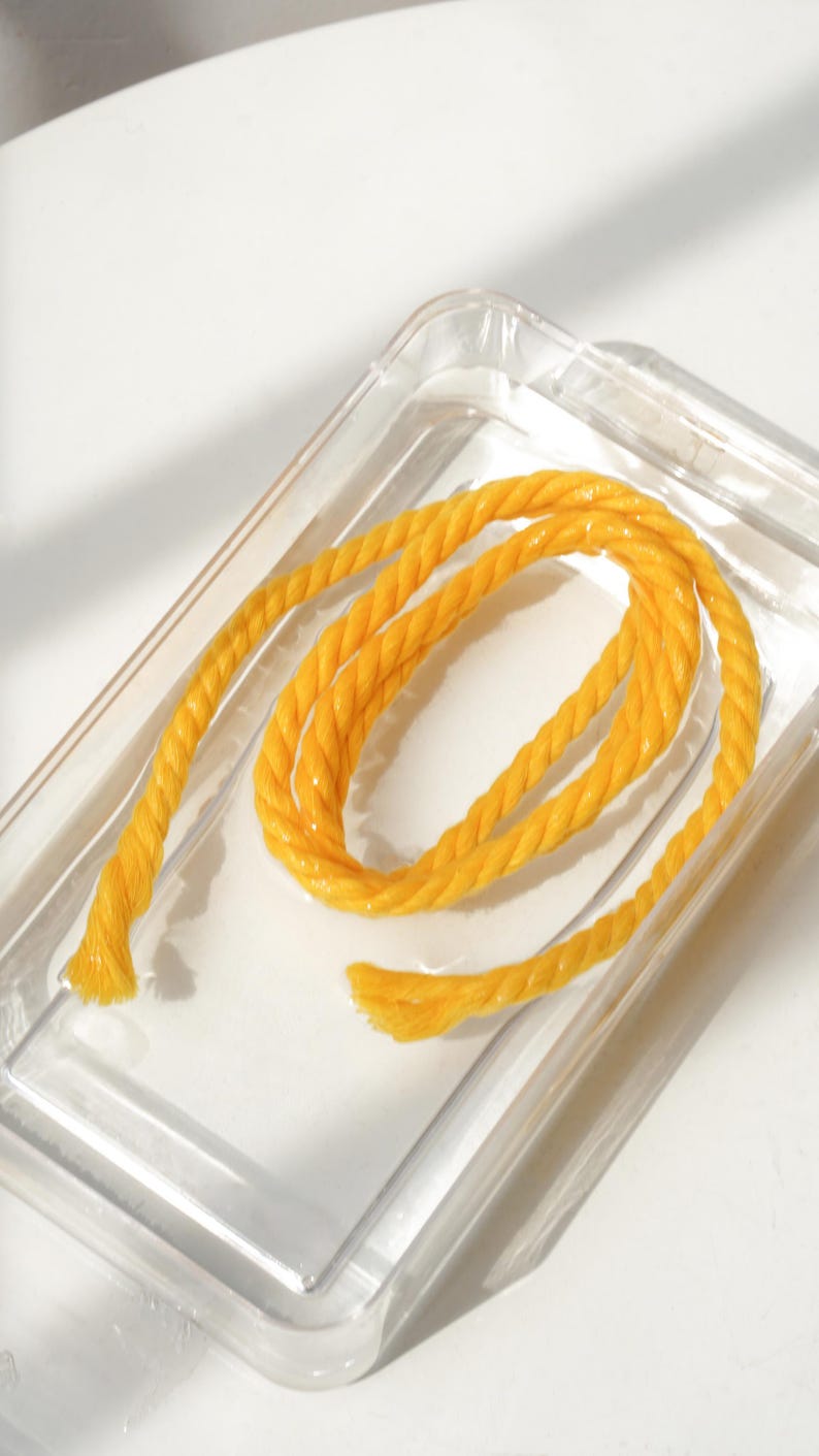 May include: A coiled length of bright yellow twisted rope with a textured surface, resting inside a clear, rectangular plastic container. The rope features a distinct braided construction with visible fibers at the frayed ends of the cord.