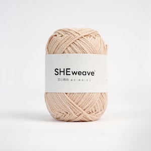 May include: A ball of beige yarn with a white label that reads "SHEweave" in black text. The label also includes text in a different language. The yarn is tightly wound and appears soft, suitable for knitting or crochet projects.