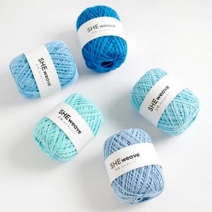 May include: Five skeins of blue yarn in varying shades, from light turquoise to deep ocean blue, are arranged on a white surface. Each skein is wrapped with a white label that reads "SHEweave". The yarn appears to be soft and suitable for knitting or crochet projects.