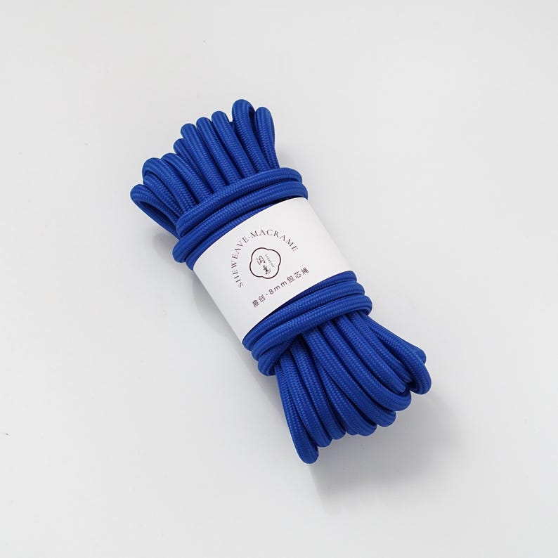 May include: A bundle of vibrant blue macrame cord, tightly wound and secured with a white label. The label features text and a logo. The cord appears to be approximately 8mm in diameter, suitable for various crafting projects.