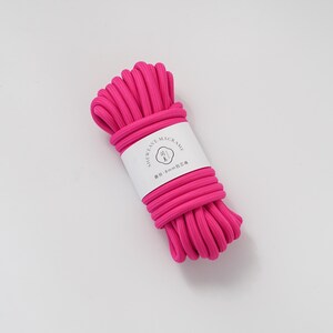 May include: A bundle of bright pink macrame cord, tightly wound and secured with a white label. The label features the text "SHEWEAVE-MACRAME" and other text. The cord appears to be approximately 8mm in diameter, suitable for various crafting projects.