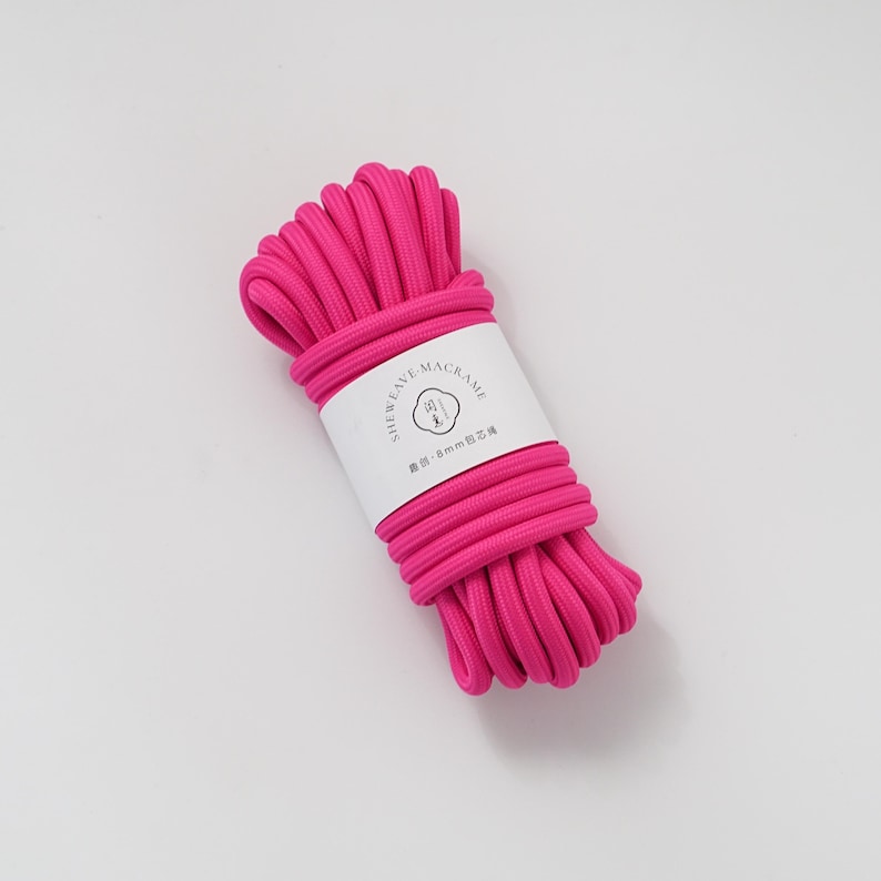 May include: A bundle of bright pink macrame cord, tightly wound and secured with a white label. The label features the text "SHEWEAVE-MACRAME" and other text. The cord appears to be approximately 8mm in diameter, suitable for various crafting projects.