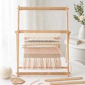 Può includere: Un telaio in legno con fili di ordito verticali bianchi e un tessuto parzialmente lavorato con fasce orizzontali di lana color crema, rosa tenue e grigio. Il pezzo tessuto presenta frange sul fondo e una navetta in legno appoggiata.