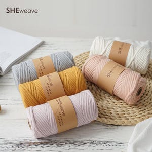 May include: Five cylindrical spools of braided cotton cord in white, gray, mustard yellow, light pink, and lavender. Each spool features a brown paper band with the text SHEweave printed in black. The cord has a thick, woven texture.