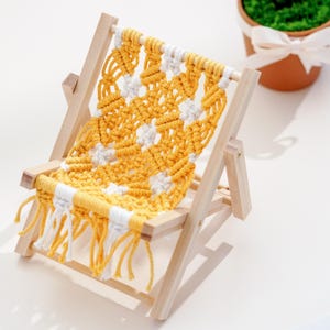 May include: Miniature wooden deck chair with a macrame seat. The seat features a yellow and white woven pattern with fringe. The chair has a natural wood frame and is designed for decorative purposes.
