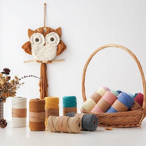 Puede incluir: Un colgante de pared de macramé con forma de búho en tonos marrón y crema, sobre una colección de coloridas bobinas de hilo. El hilo, en tonos como blanco, amarillo, turquesa y rosa, está empaquetado con etiquetas de papel marrón. También se ve una cesta de mimbre.