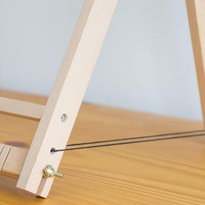 May include: A wooden weaving loom with a light wood finish. The loom has a series of vertical slots and a brass knob. Black strings are strung across the frame. The loom is set on a wooden surface.