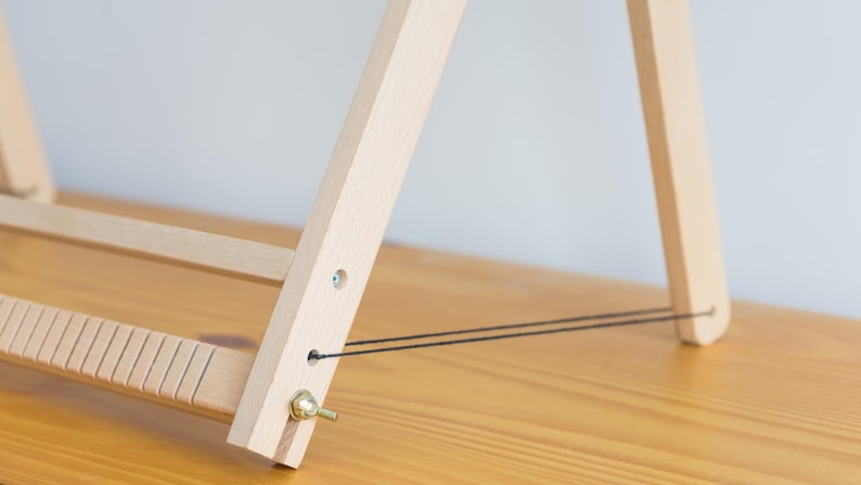 May include: A wooden weaving loom with a light wood finish. The loom has a series of vertical slots and a brass knob. Black strings are strung across the frame. The loom is set on a wooden surface.