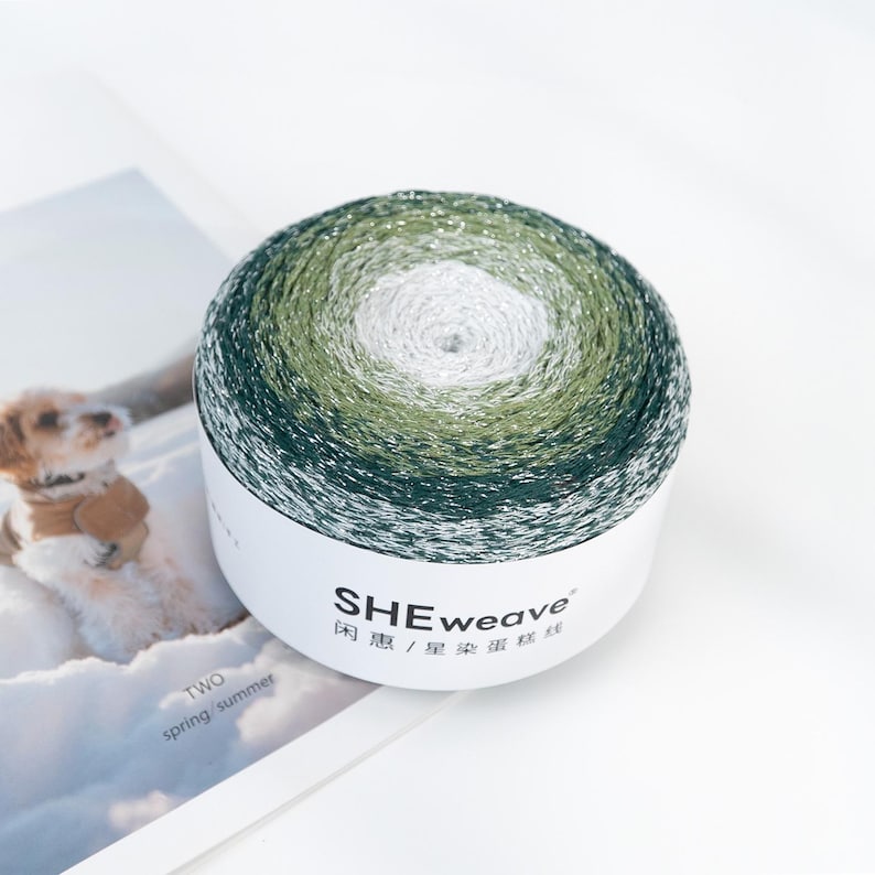 May include: A round skein of yarn with a gradient of colors, transitioning from dark green to light green and white. The yarn is wrapped around a white cardboard tube with the brand name "SHEweave" printed on it. The text "TWO spring/summer" is also visible.