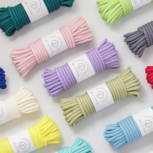Pode incluir: Uma variedade de rolos de corda de macramé coloridos em vários tons, incluindo rosa, verde, azul, amarelo e cinza. Cada rolo é embrulhado com um rótulo branco com um logotipo. As cordas estão bem organizadas, mostrando uma variedade de materiais de artesanato.