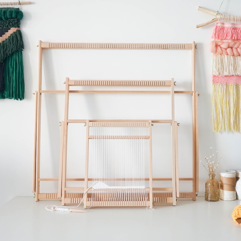 May include: Three rectangular wooden weaving looms of varying sizes nested together. Each loom features a light-colored wood frame with evenly spaced notches along the top and bottom bars. The smallest loom is partially warped with white yarn.